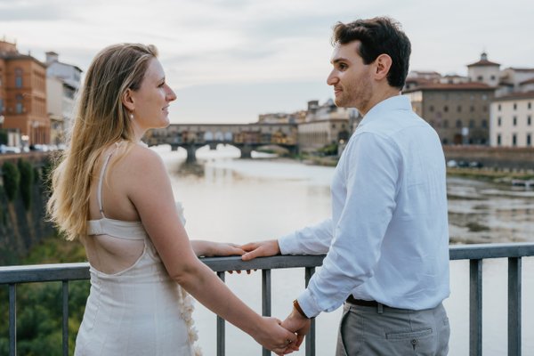 Couple holding hands by the Arno River with historic Florence bridges and buildings behind them in daylight
