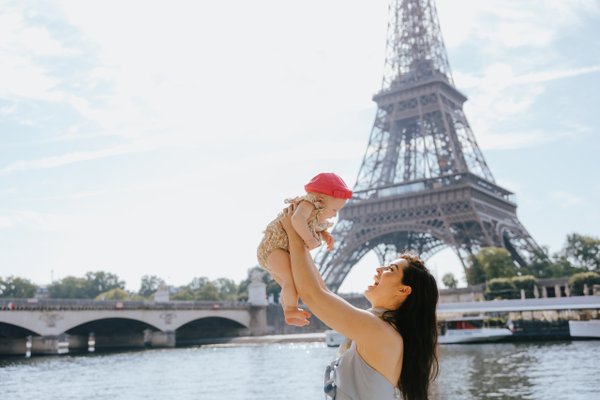Mother holding toddler in red cap by Eiffel Tower and Seine River on bright daytime