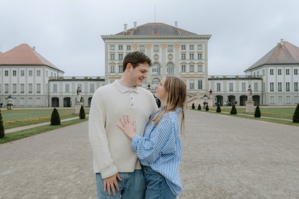 Couple smiling together in front of Nymphenburg Palace's ornate baroque buildings on an overcast day