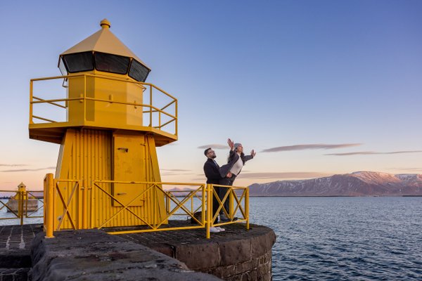 Couple posing by a yellow lighthouse at dusk with mountain and water views in Reykjavík