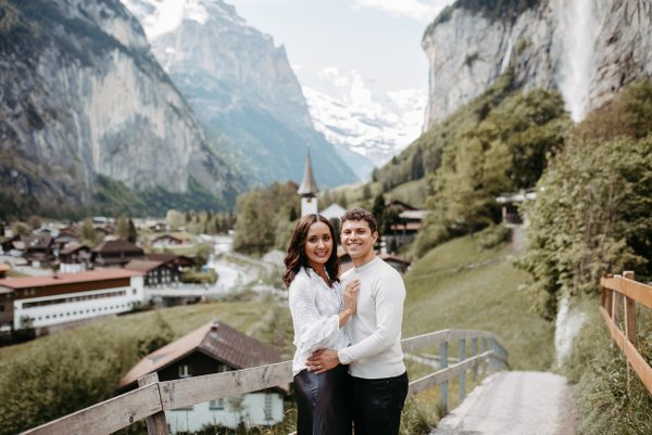 Couple posing on a mountain balcony overlooking an alpine valley with cliffs, a church spire, and snow-capped peaks in Interlaken