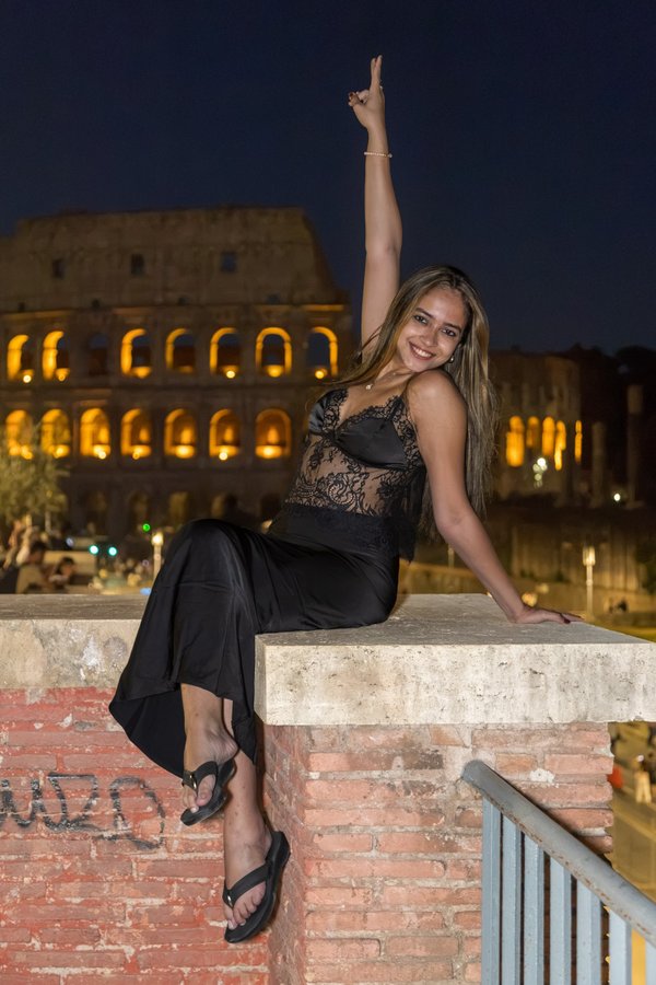 Woman in black outfit posing on ancient wall with illuminated Colosseum at night in Rome