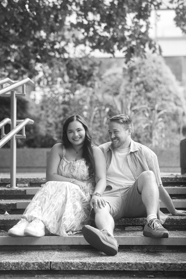 Couple sits on outdoor steps beneath tree foliage in black and white portrait