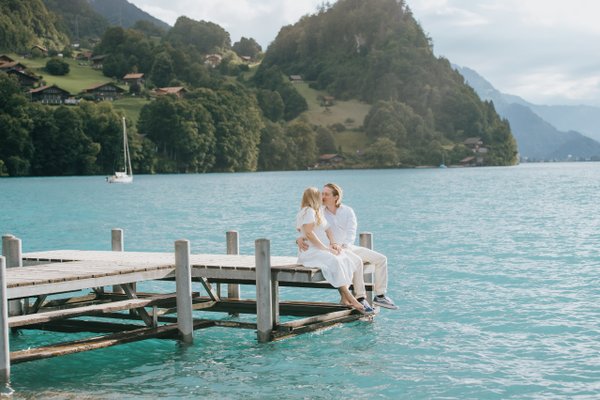 Couple in white sitting on wooden pier over turquoise alpine lake, forested mountains rising behind, daytime