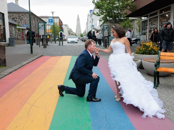 Groom kneeling on rainbow crosswalk toward bride in white dress, Hallgrímskirkja church visible behind in Reykjavík