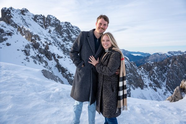 Smiling couple in winter coats showing off an engagement ring against snowy alpine peaks at sunny midday