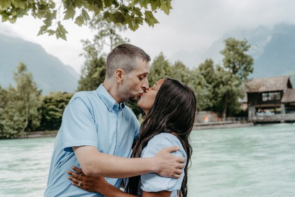 Couple kissing by turquoise river with alpine mountains and boathouse in background during daytime