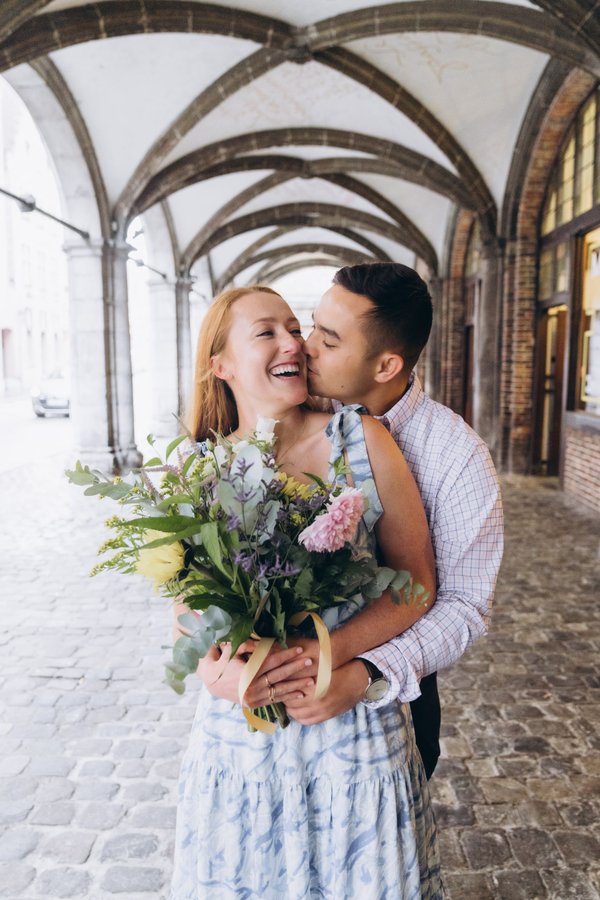 Couple embracing under arched stone corridor holding fresh flowers bouquet during daytime in Bruges