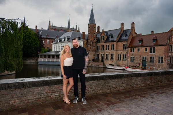 Couple posing on brick waterfront with medieval towers and canal buildings behind them under cloudy sky