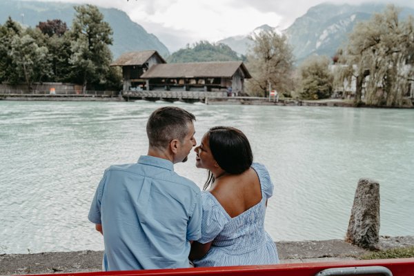 Couple sitting by turquoise river with boathouse and alpine mountains under cloudy sky in Interlaken