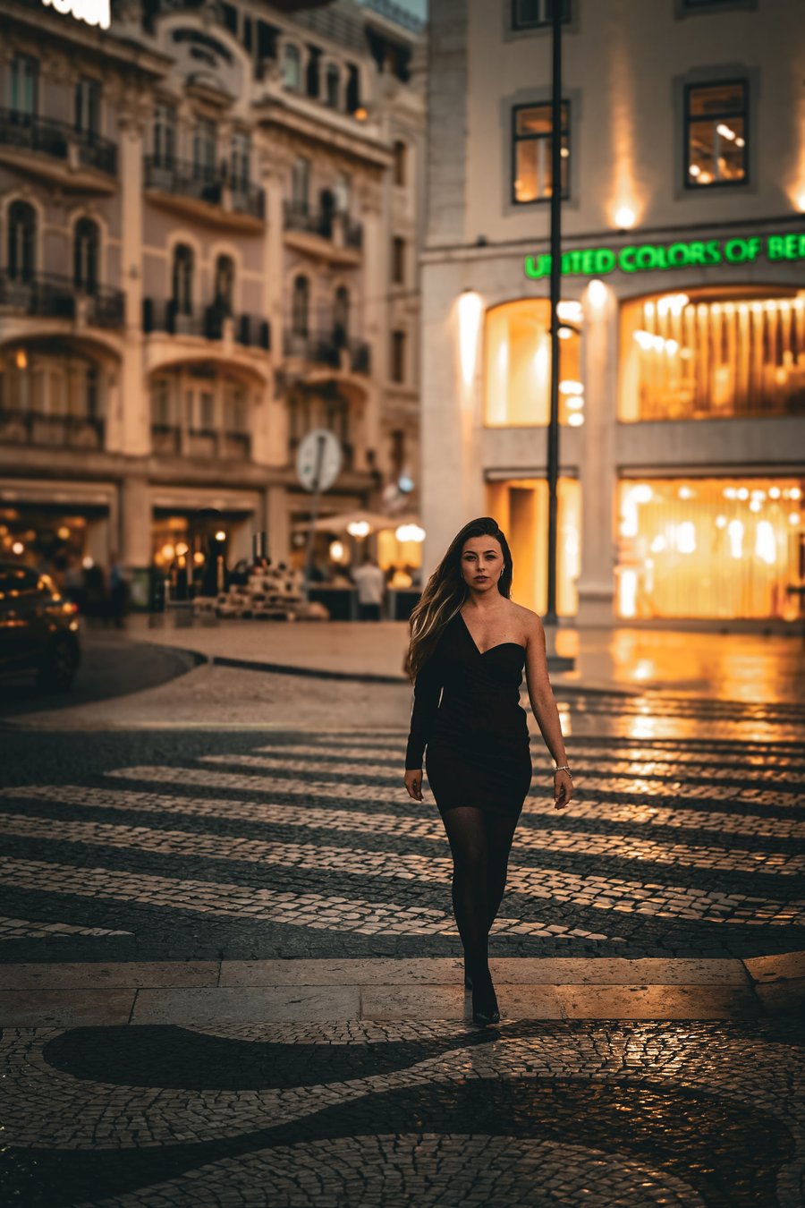 Woman in black dress walking through illuminated Cascais old town street at evening with historic architecture and storefronts