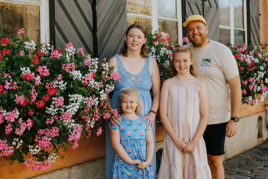 Family of four smiling beside vibrant pink and red flowers against a rustic building in Helsinki city center