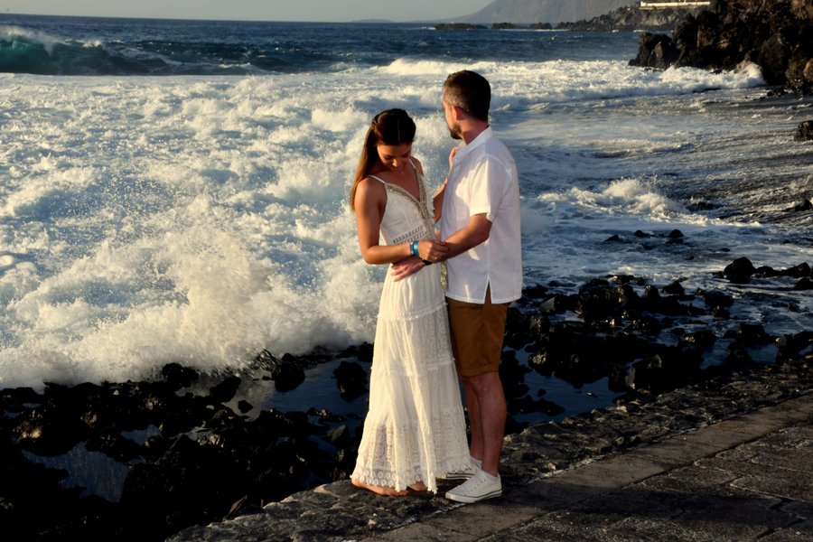 Couple in white holding hands on rocky beach at sunset with crashing waves