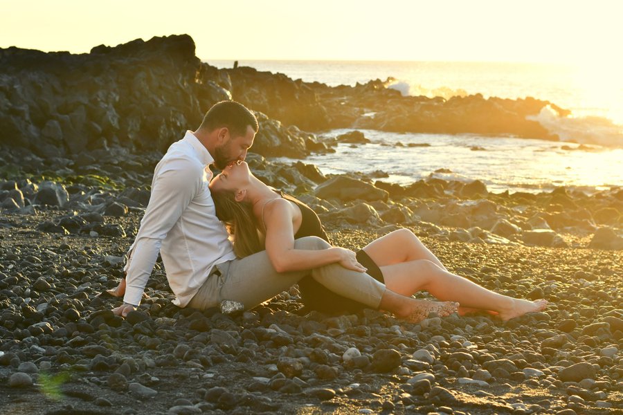 Couple embracing on rocky beach at sunset, golden hour light over ocean and coastal cliffs