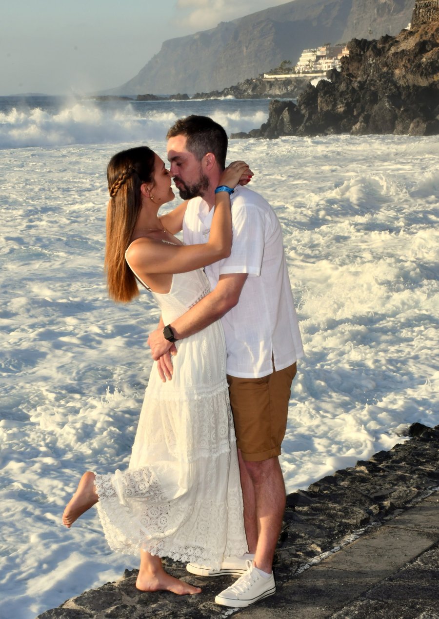 Couple kissing in white lace dress and shirt on rocky beach with waves and coastal cliffs in golden sunlight