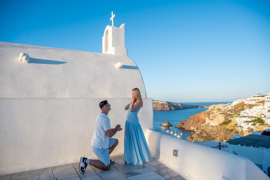 Man kneeling to propose to woman in blue dress at white church in Santorini, overlooking caldera at sunset