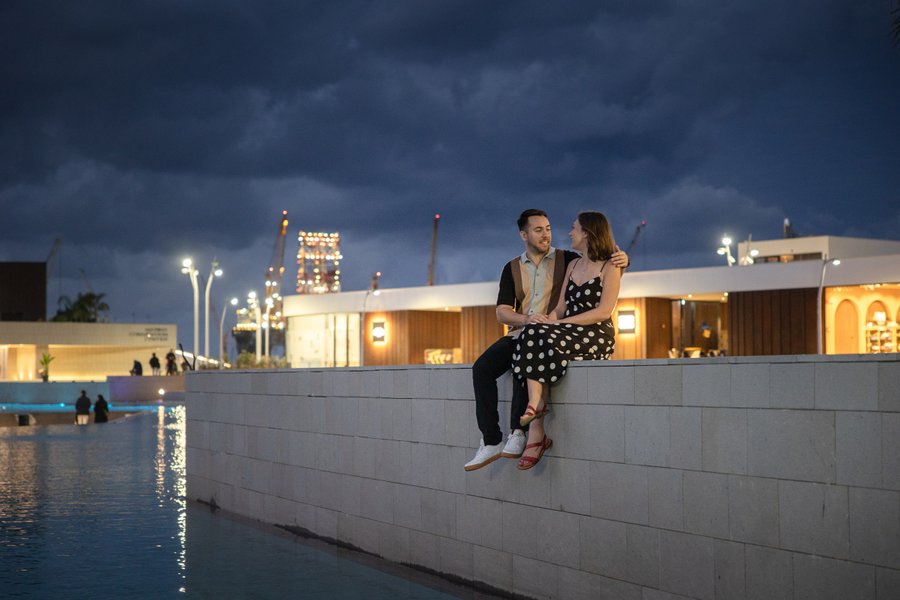 Couple sitting on waterfront wall at dusk with illuminated modern architecture and Eiffel Tower replica in background