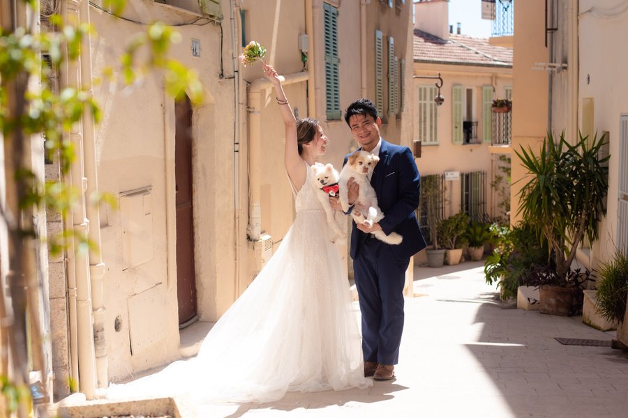 Bride and groom with white dog in sunny old town alley, holding greenery, classic European architecture