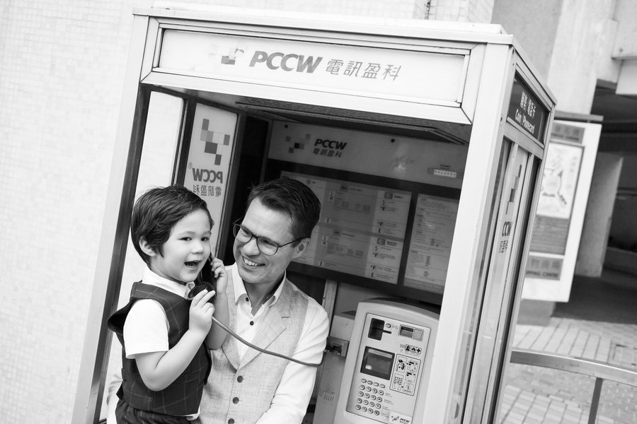 Father and young son smiling inside a vintage Hong Kong phone booth, black and white portrait