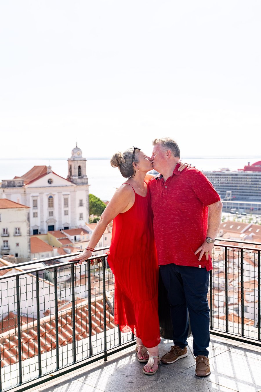Couple kissing on a sunny rooftop terrace overlooking Alfama's white church and red-tiled roofs in Lisbon