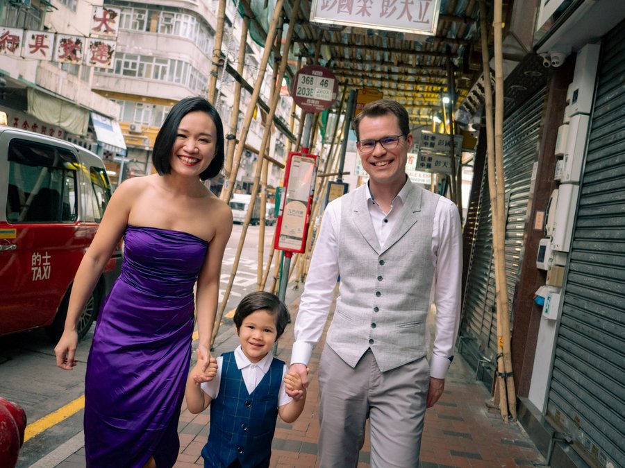Family of three walking hand-in-hand through a narrow Hong Kong street with traditional shopfronts and Chinese signage
