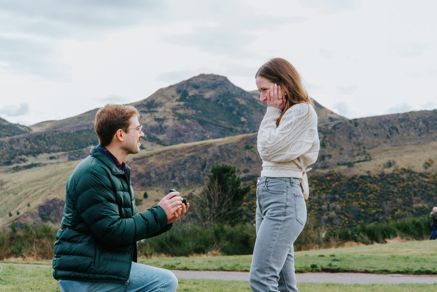 Couple in casual outfits on grassy hillside with dramatic mountain backdrop under cloudy sky