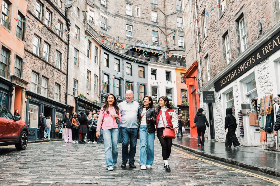 Family of four walking through Edinburgh's wet cobblestone old town street lined with historic stone buildings and shops
