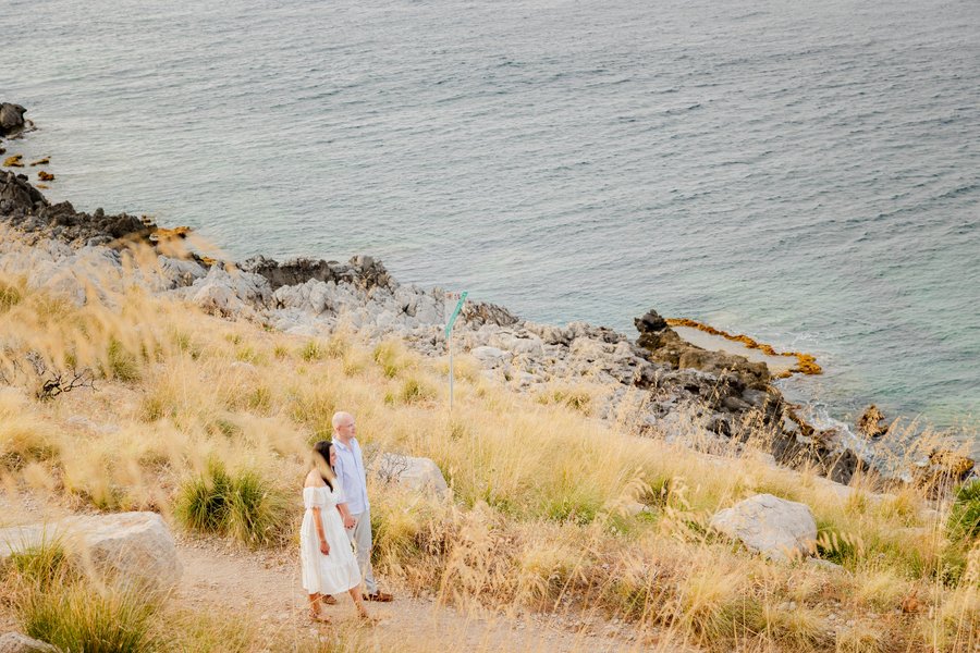 Couple standing on dry rocky hillside overlooking turquoise Mediterranean water at golden hour