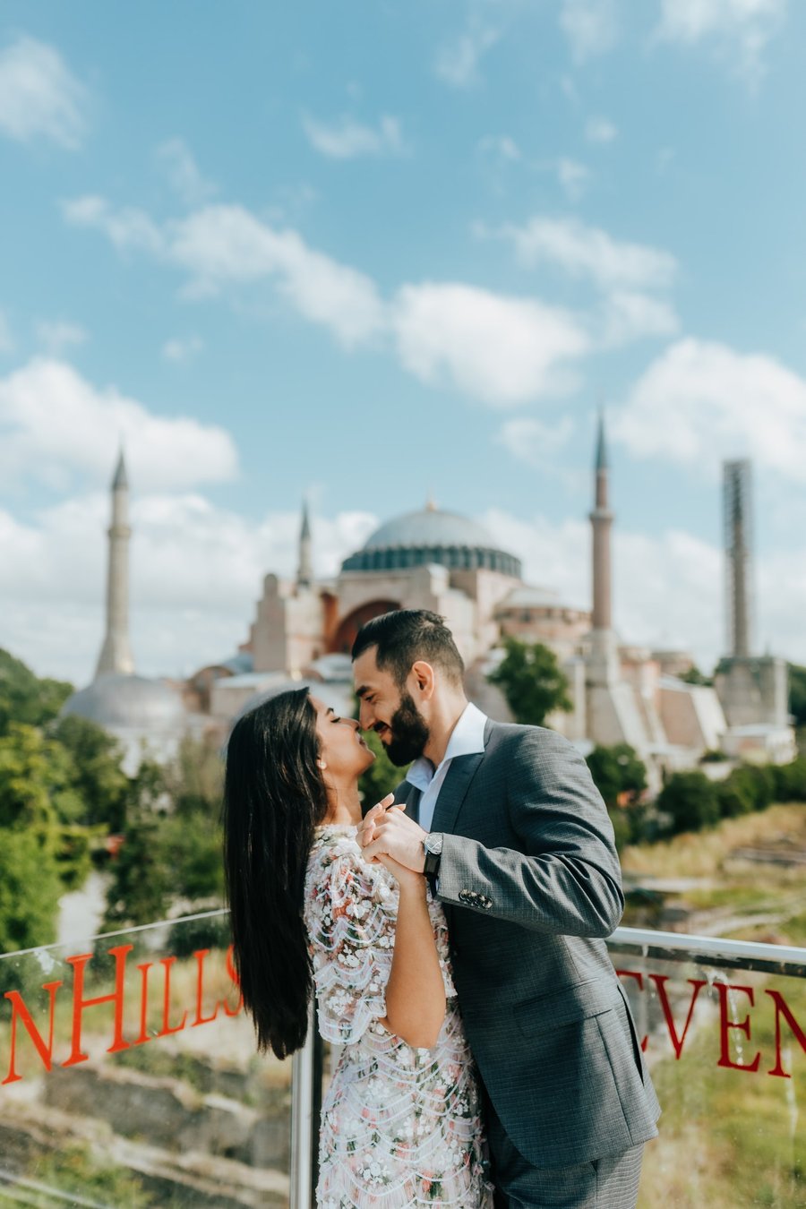 Couple in formal attire kissing with Hagia Sophia and Blue Mosque blurred behind them under blue sky
