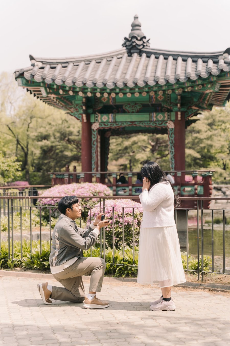 Man proposing to woman in white áo dài at Asian pavilion with pink flowers and green gardens in daylight