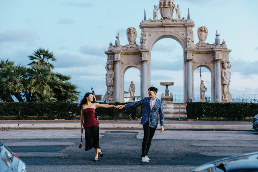couple holding hands on street with ornate stone archway and palm trees under blue sky
