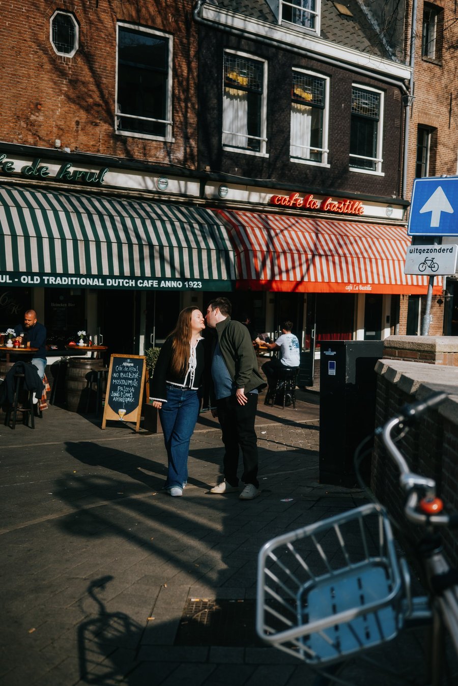 Couple embracing outside a traditional Dutch café on a sunny street in Utrecht, with bicycles and historic brick buildings nearby