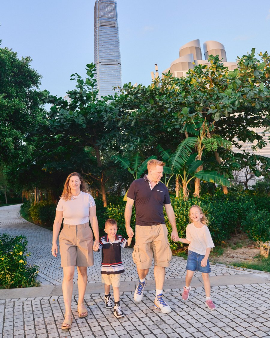 Family of four walking through lush garden with modern skyscraper and curved building in background during golden hour