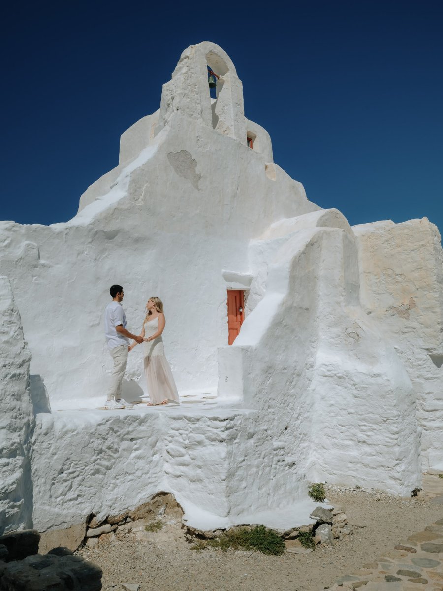 Couple in light clothing standing before whitewashed Paraportiani Church under clear blue sky in bright daylight