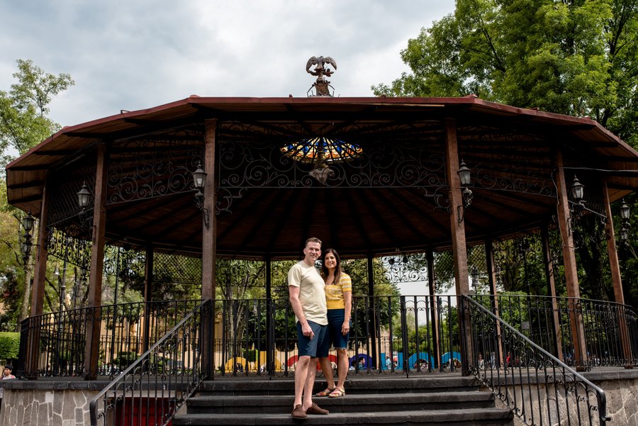 Couple posing at ornate wooden gazebo with decorative ironwork in lush green park setting