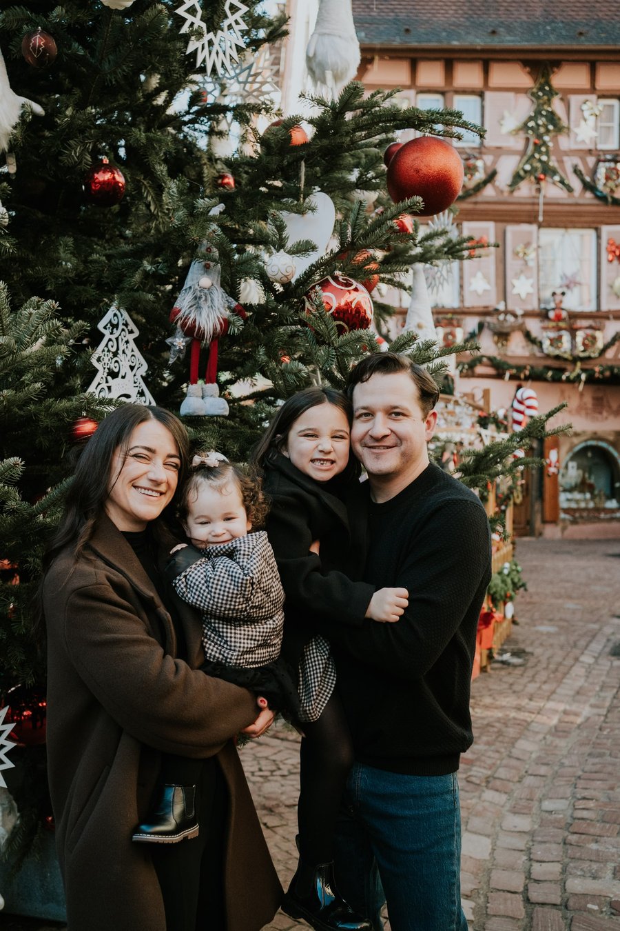 Family of four smiling among decorated Christmas trees in a Colmar street during daytime