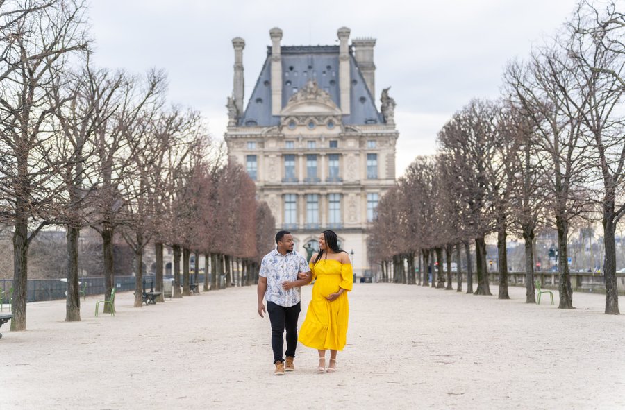 Couple walking arm-in-arm down a tree-lined pathway toward a grand Parisian palace in daylight