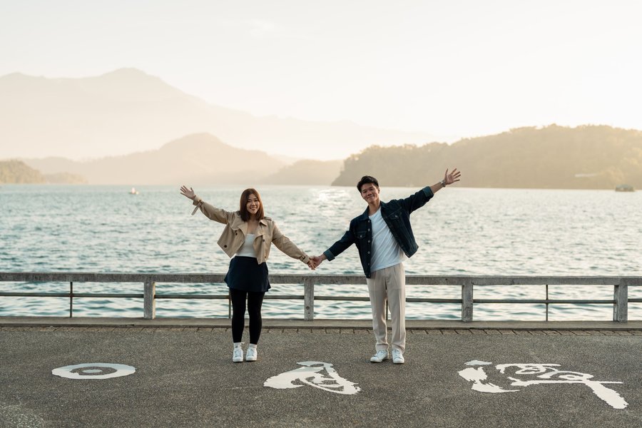 Couple holding hands with arms raised at lakeside promenade, mountains and water in background during overcast day