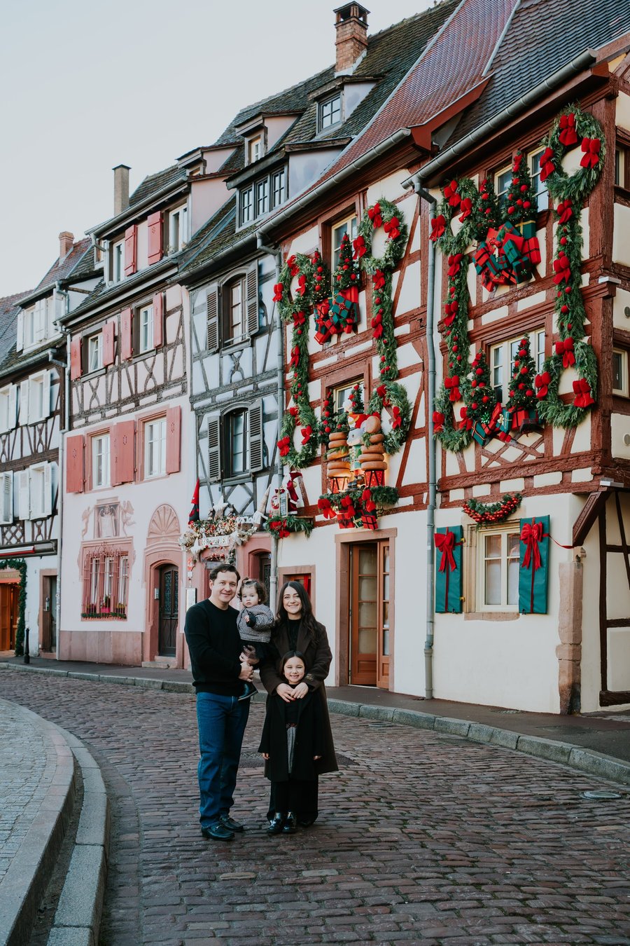 Family of four standing on cobblestone street lined with half-timbered buildings decorated with red Christmas garland