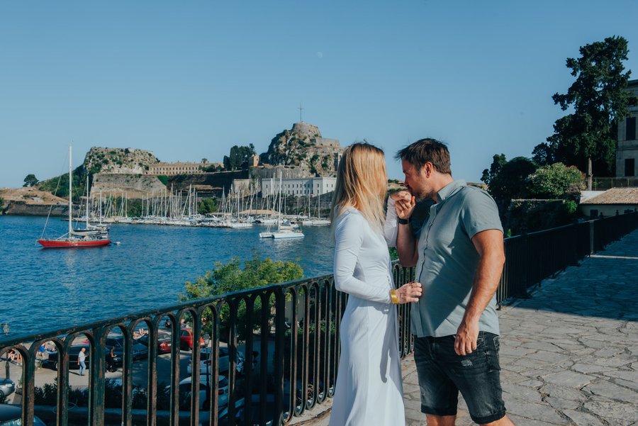 Couple enjoying clear blue skies overlooking a Mediterranean harbor with sailboats and historic fortress in background