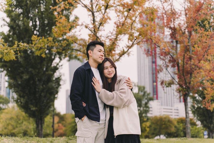 Couple posing together among autumn trees with city skyscrapers in the background during fall season