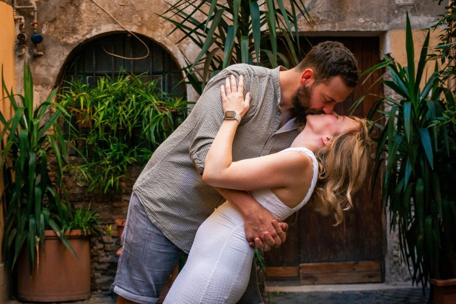 Couple kissing in a lush courtyard surrounded by potted green plants and rustic terracotta walls in Rome