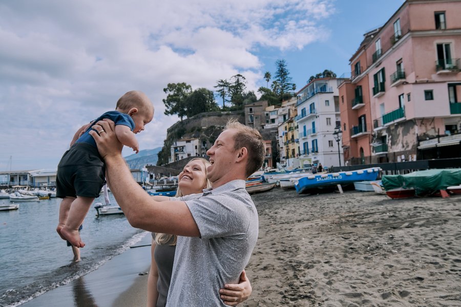 Family playing on a sandy beach in Sorrento with colorful waterfront buildings and boats in bright daylight