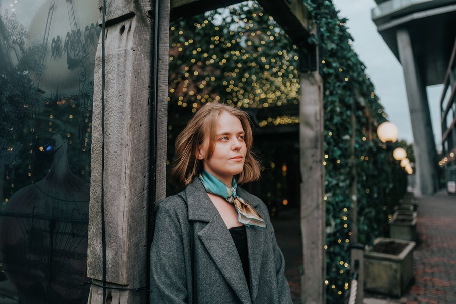 Solo woman in gray blazer standing in ivy-covered alleyway at dusk with golden bokeh lights