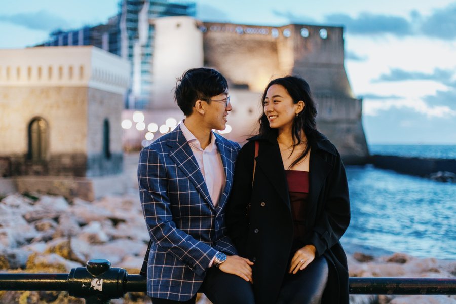 Couple smiling at waterfront with historic castle and harbor buildings at dusk in Naples