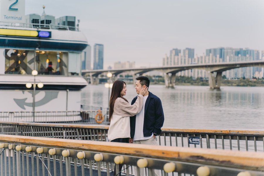 Couple on a waterfront pier with a ferry and Seoul skyline reflected in calm water behind them
