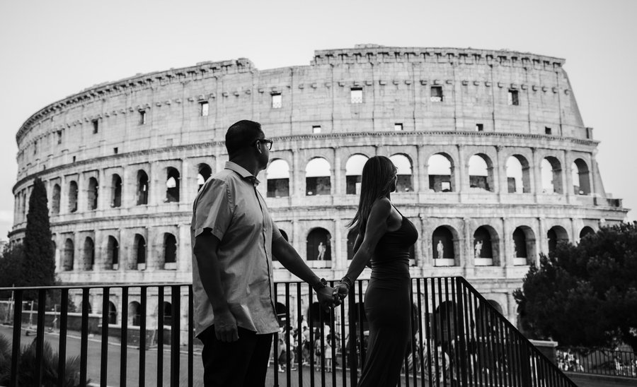 Couple holding hands at a railing with the ancient Colosseum in black and white behind them