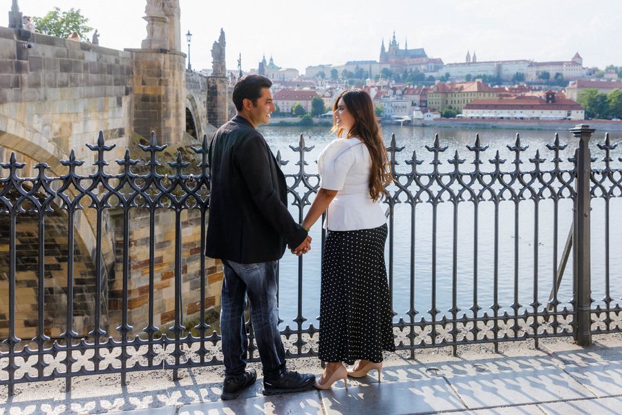 Couple holding hands by ornate railing overlooking Prague's Vltava River and historic cityscape at daytime