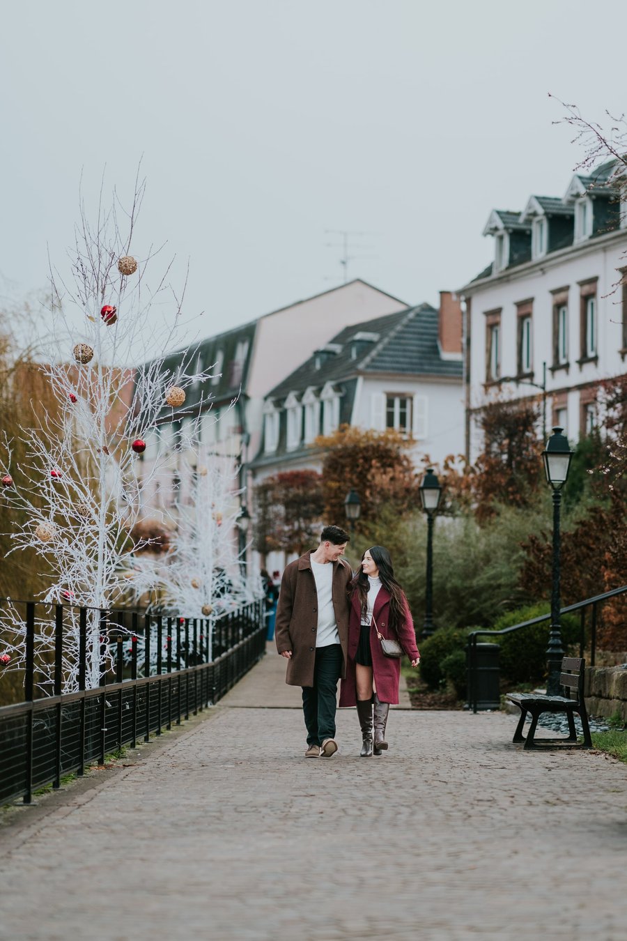 Couple walking through festive European street with white ornament-decorated tree and historic architecture in Colmar