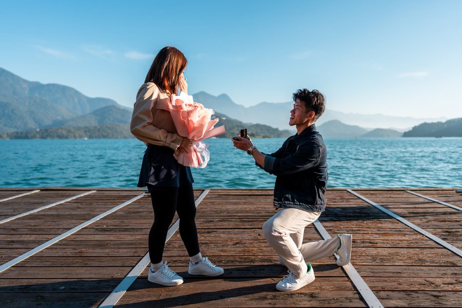 Man proposing to woman holding pink flowers on wooden dock overlooking mountains and turquoise lake